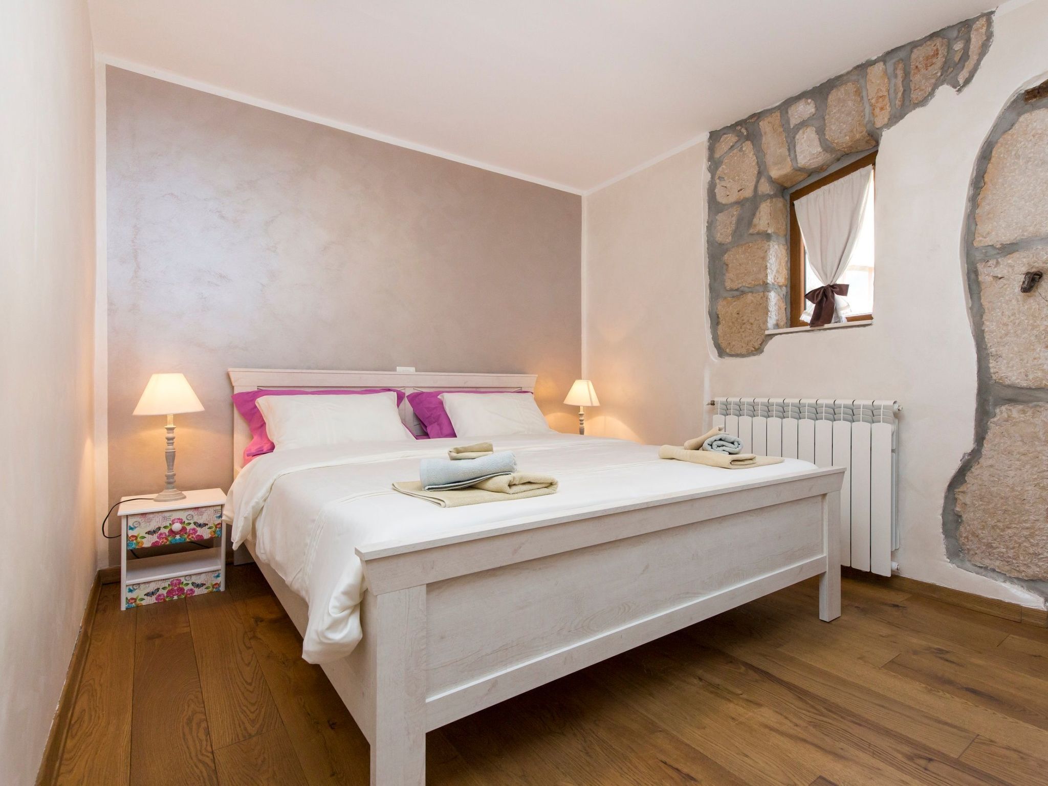 A bedroom with a bed, bedside lamps, a radiator, and a stone wall.