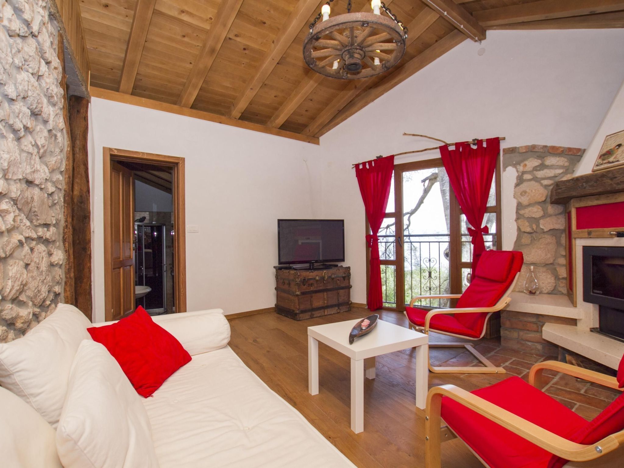 A cozy living room with a stone wall, wooden ceiling, and a fireplace. There is a white sofa with red cushions, a red armchair, and a television on a wooden cabinet.