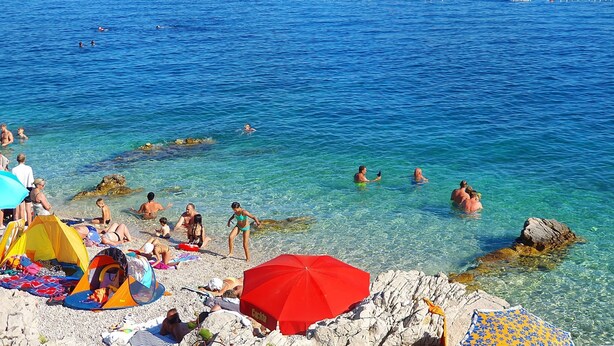 Una spiaggia con acqua cristallina, persone che nuotano e si rilassano al sole, e una barca all'orizzonte.