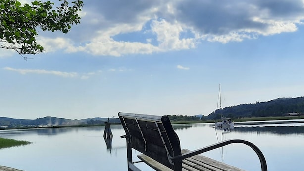 Um banco à beira de um lago tranquilo, com um veleiro ao longe e colinas ao fundo.