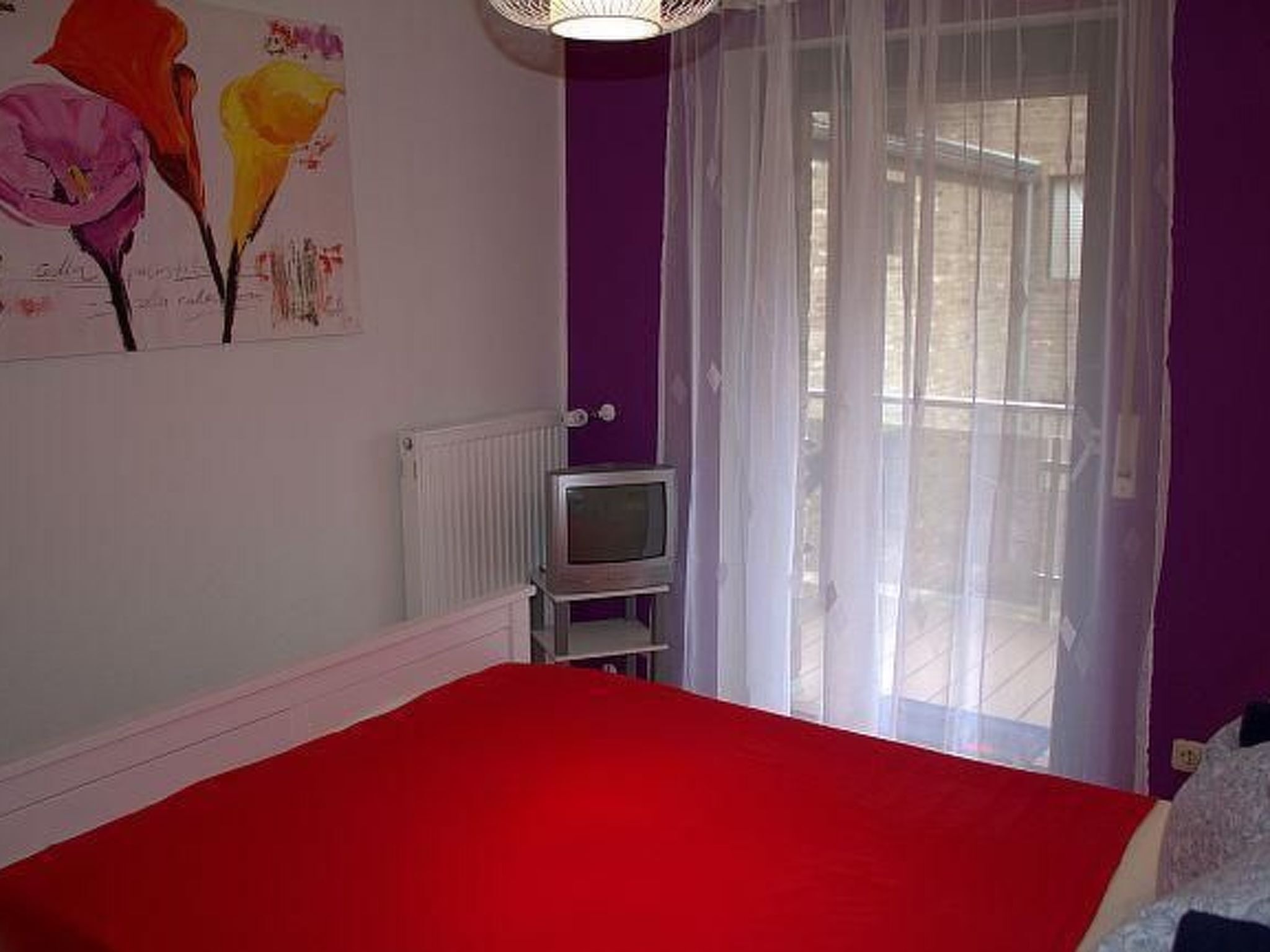 Un dormitorio con una colcha roja, un televisor sobre un mueble bajo y una ventana con cortinas traslúcidas.