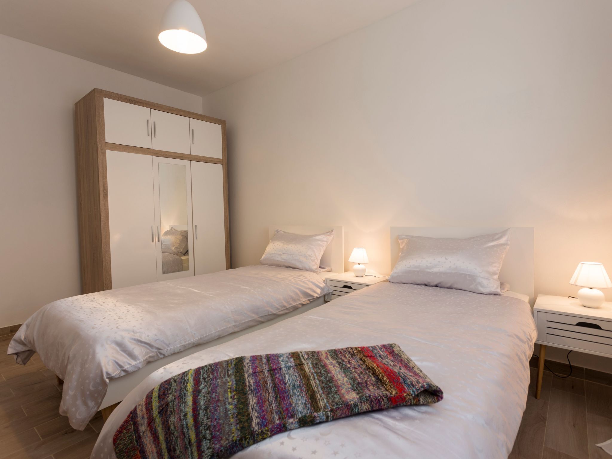 Uma cama de casal com um cobertor colorido, uma mesa de cabeceira com abajur e um guarda-roupa com portas espelhadas.