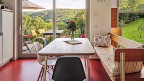 A room with a dining table, chairs, a bench, a hanging light fixture, and a large window with a view of a forested landscape. - Moderner Holzhausneubau in der Eifel by Interhome (Berlingen)