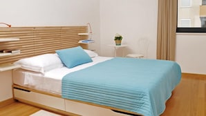 Ein modernes Schlafzimmer mit einem Holz-Kopfteil, einem Bett mit blauer Steppdecke, einem Nachttisch mit Lampe, einem kleinen Tisch mit Pflanze und einem Fenster mit Vorhängen.
