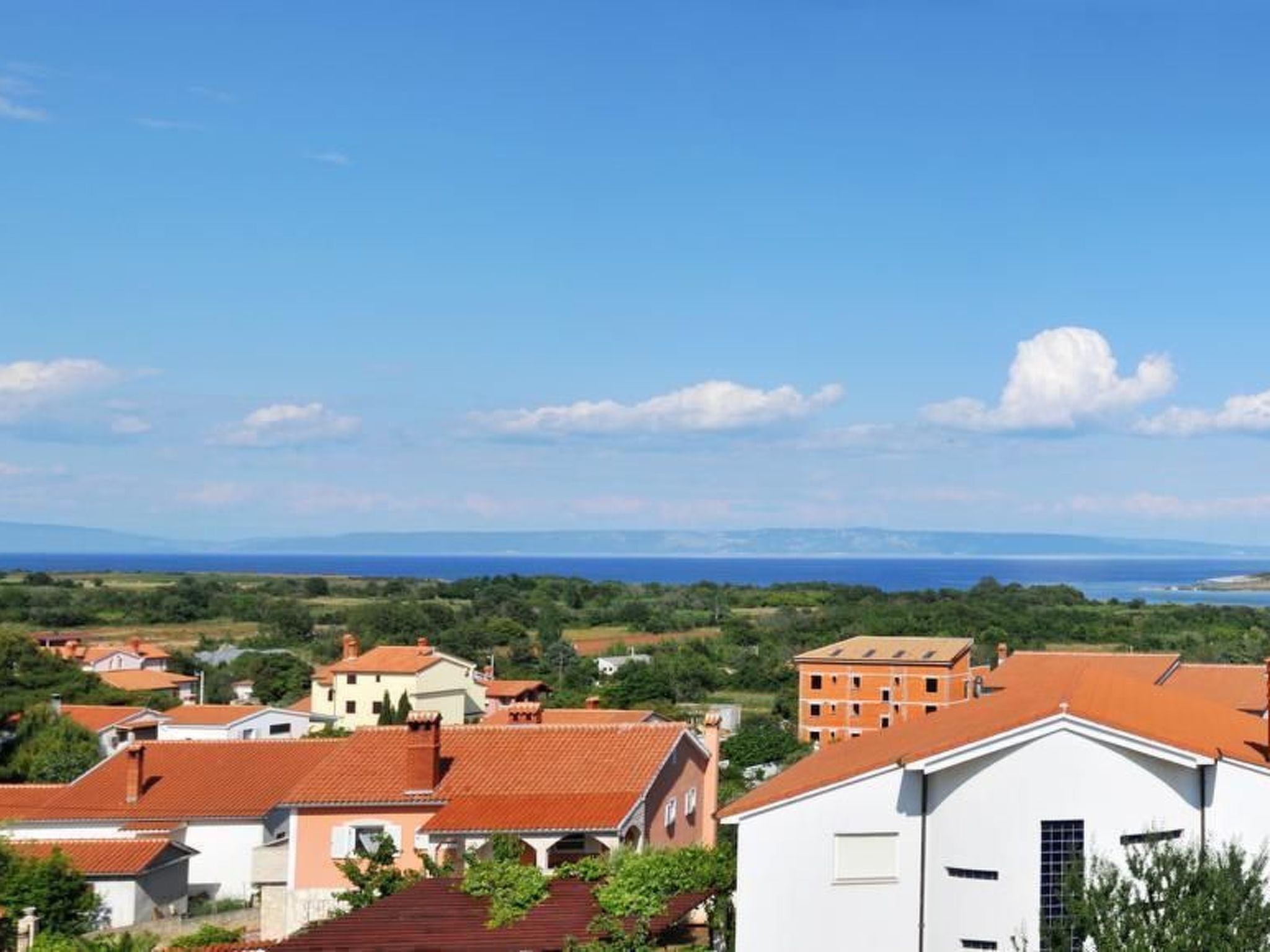 A coastal town with terracotta-roofed buildings, a white building with a large window, and a view of the sea and distant mountains.