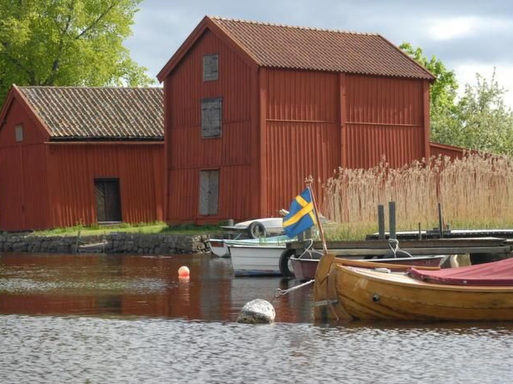 A red wooden building with a sloped roof, a dock with boats, and a Swedish flag.