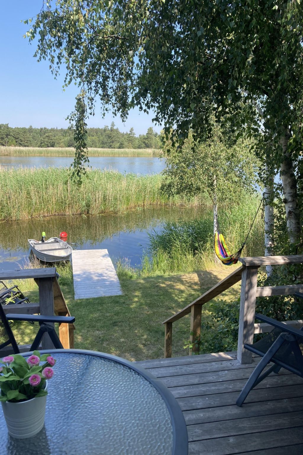 Una terraza de madera con una mesa y sillas de vidrio, con vista a un lago tranquilo con un muelle y una hamaca.