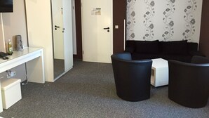 Habitación de hotel con escritorio, dos sillas negras, un sofá, un televisor y una puerta con un cartel.