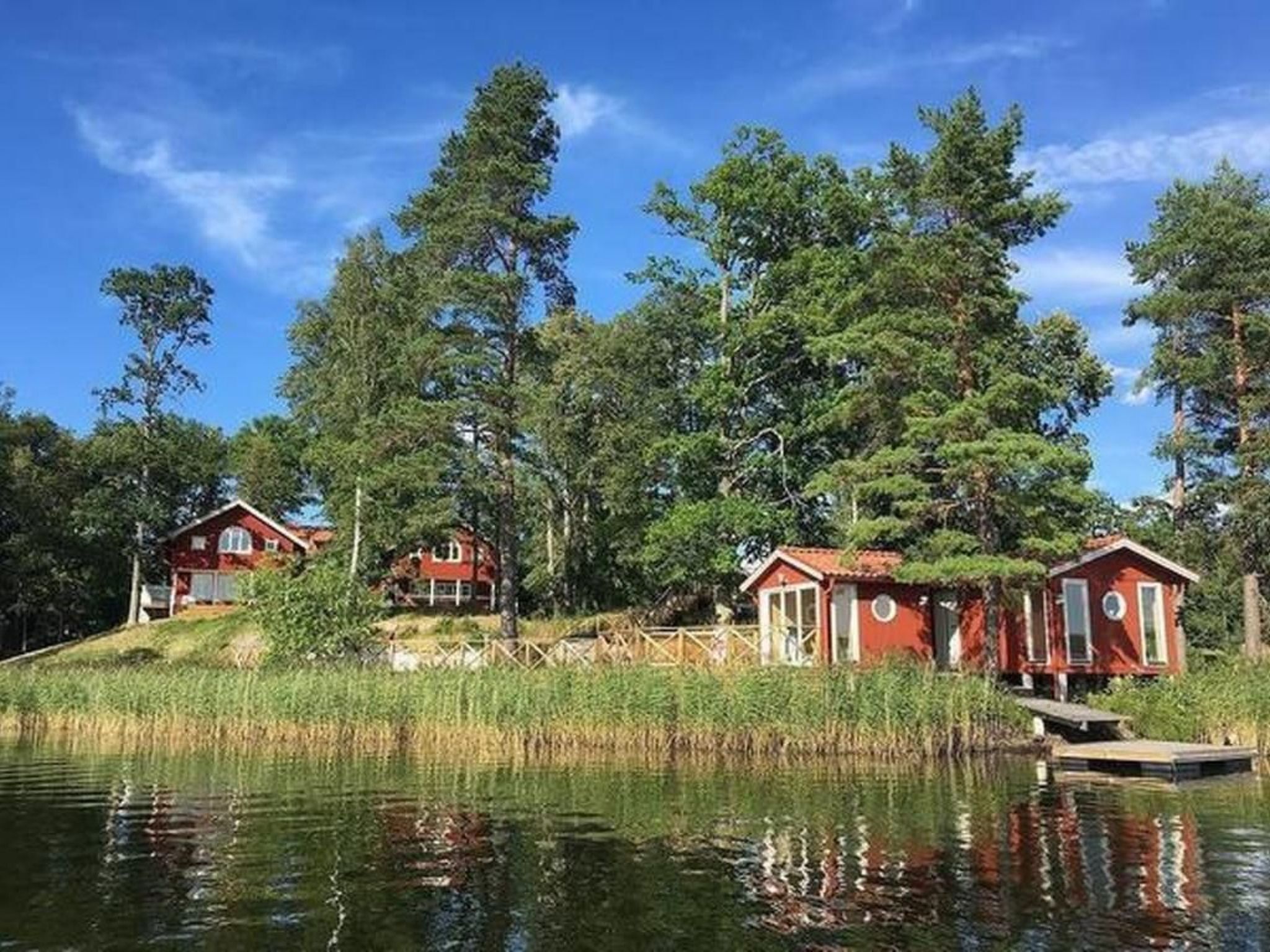 Une propriété au bord du lac, comprenant trois cabanes rouges, chacune dotée d’un petit quai, et entourée d’une forêt dense et d’un ciel bleu clair.