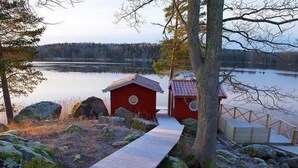 Ein hölzerner Weg führt zu einer roten Holzhütte am See, umgeben von Felsen und Bäumen.