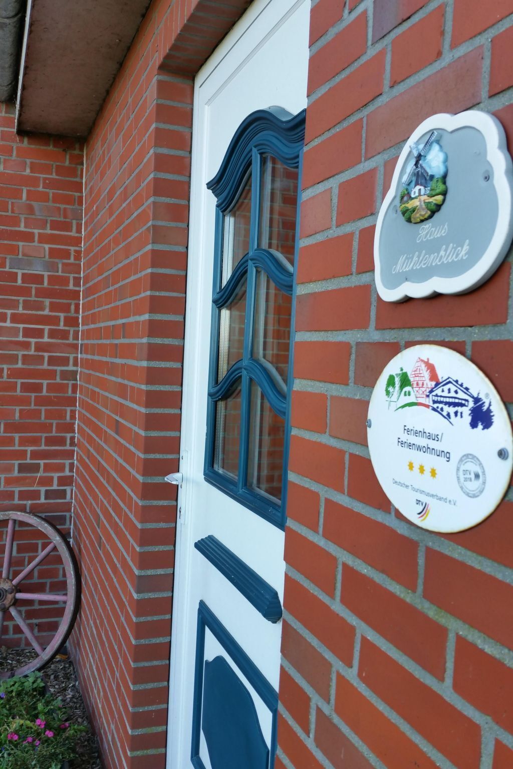 Un bâtiment en briques avec une porte blanche et des encadrements de fenêtres bleus. On peut voir deux plaques sur le mur, l’une portant l’inscription « Haus Mühlenblick » et l’autre, « Ferienhaus/Ferienwohnung », ainsi que le logo d’une association allemande de tourisme.
