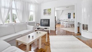 A spacious living room with a white sofa, a coffee table, and a television. There is a dining area with a table and chairs visible in the background. - Rosenhill by Interhome (Pauliström)