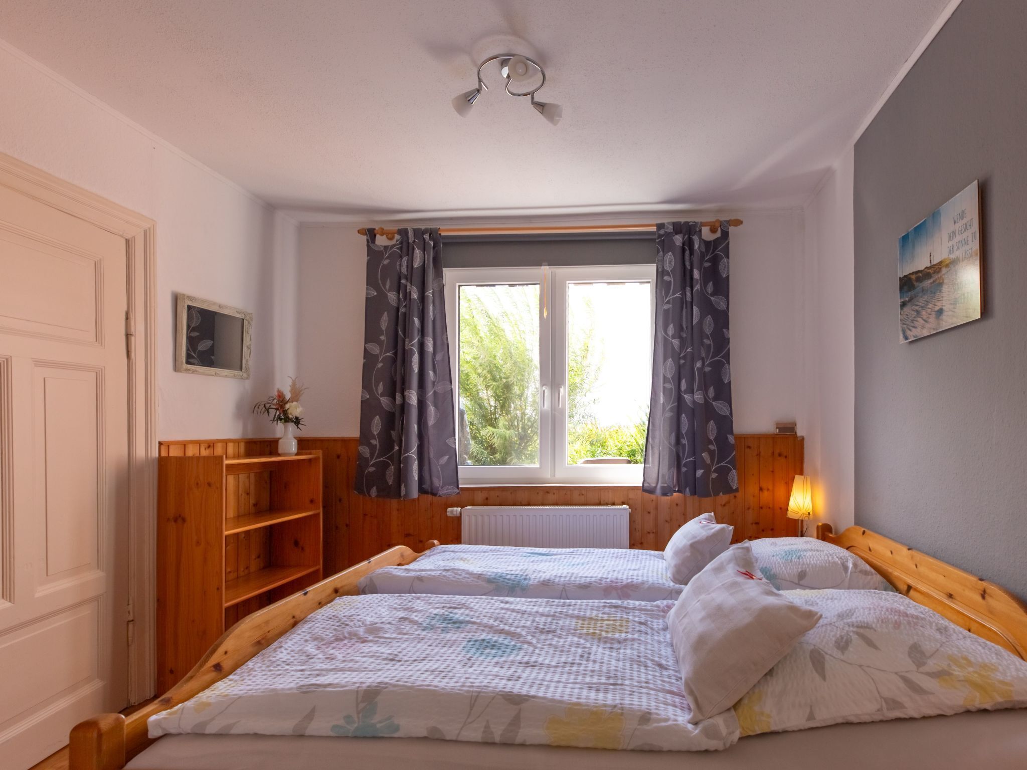 Un dormitorio con dos camas, una ventana con cortinas, un radiador, un estante de madera, una lámpara de noche y un cuadro en la pared.