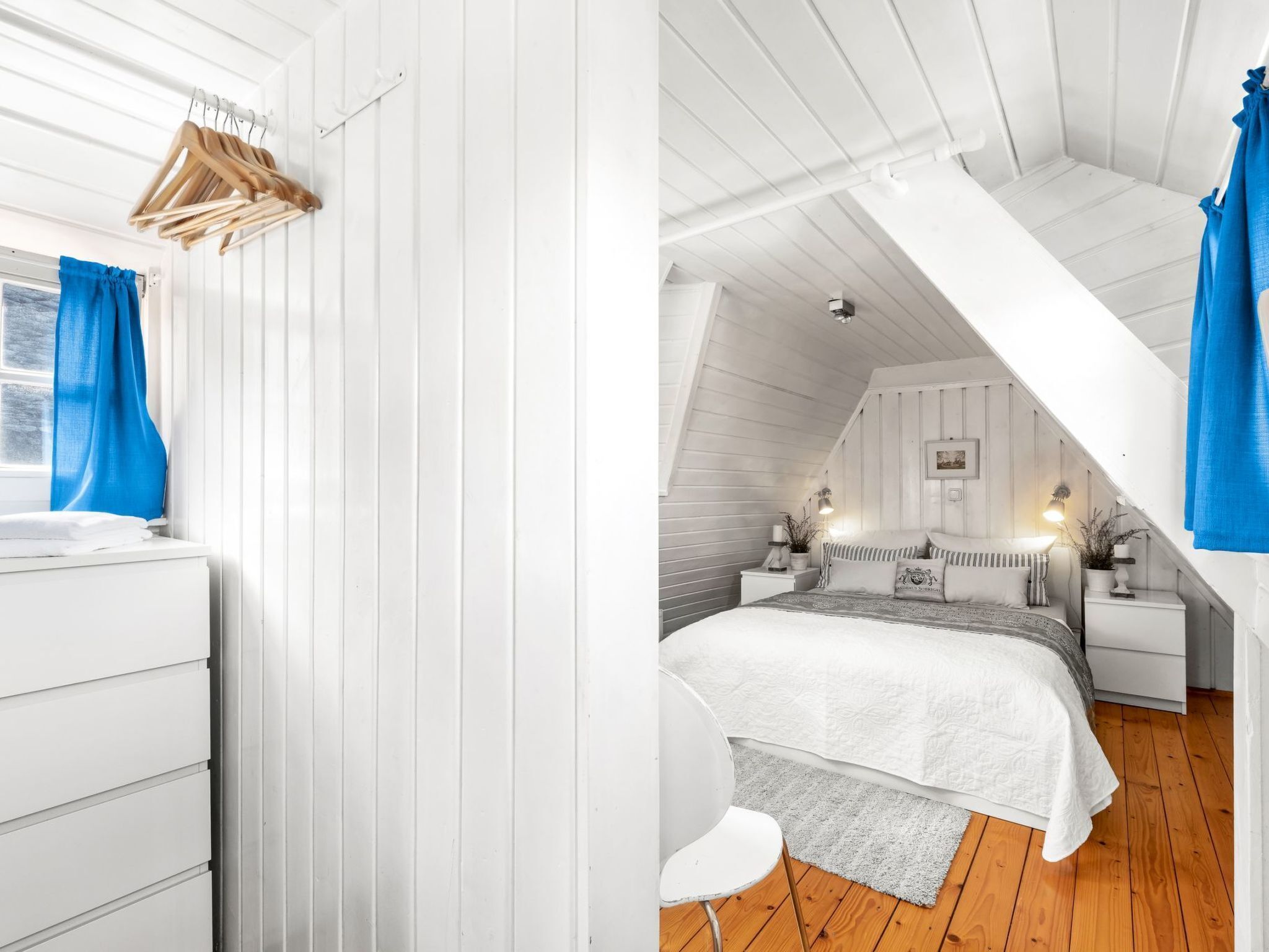 Een slaapkamer met een schuine balkenlaag, een bed met een witte dekbedovertrek, een nachtkastje met een lamp en een raam met blauwe gordijnen.