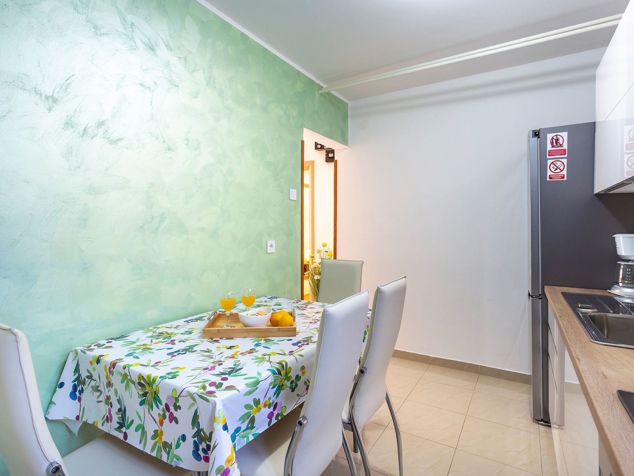 A small kitchen with a dining area. There is a table with a floral tablecloth, two chairs, and a tray with oranges and glasses. A refrigerator is visible on the right side.