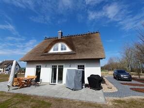 Une maison avec un toit de chaume, une terrasse avec une table et des chaises, et une voiture garée à proximité.
