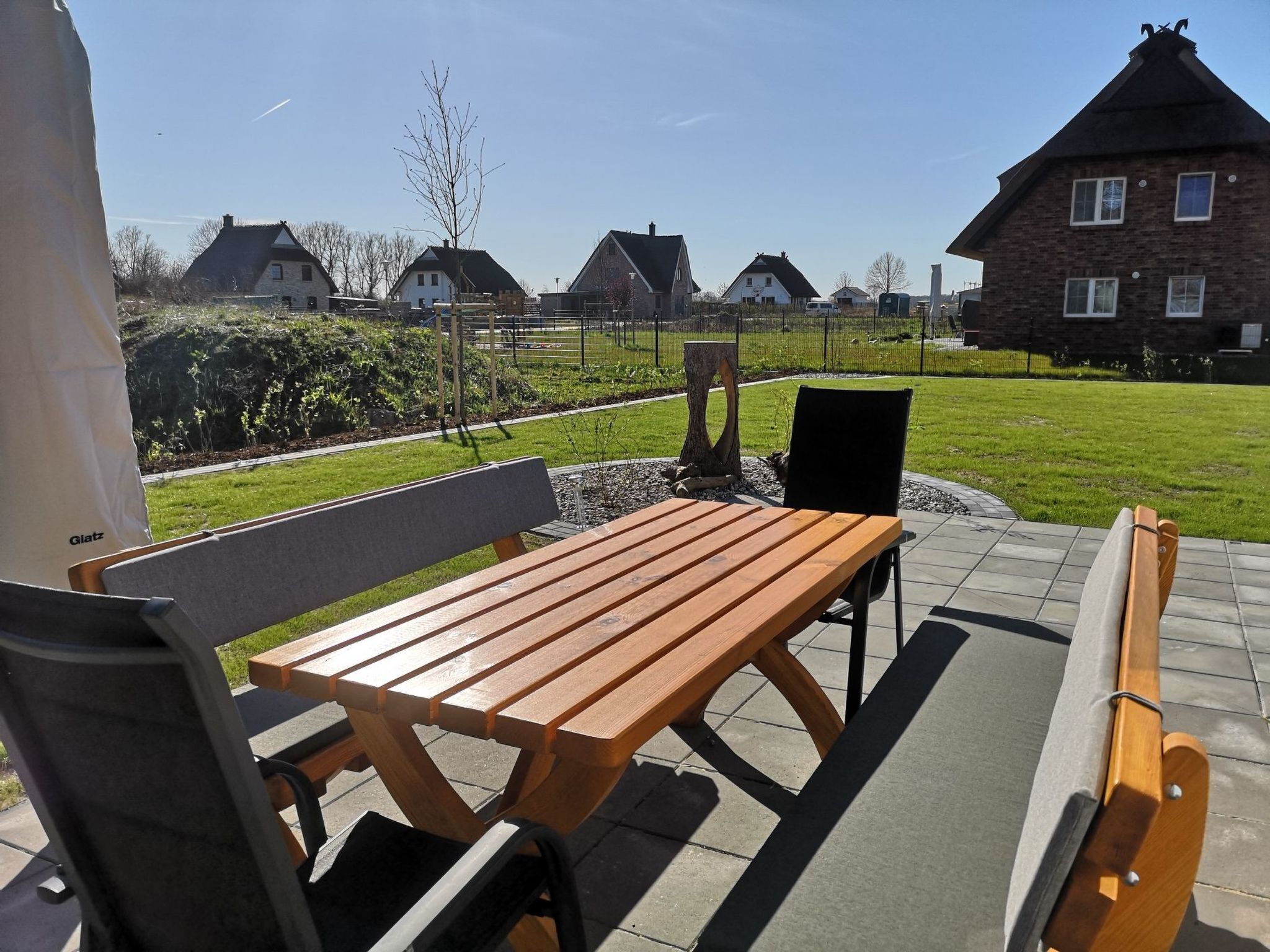Een terras buiten met een houten tafel en bankjes, omringd door een verzorgd gazon en een stenen muur.