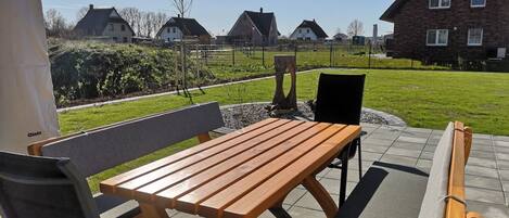 Een terras buiten met een houten tafel en bankjes, omringd door een verzorgd gazon en een stenen muur.