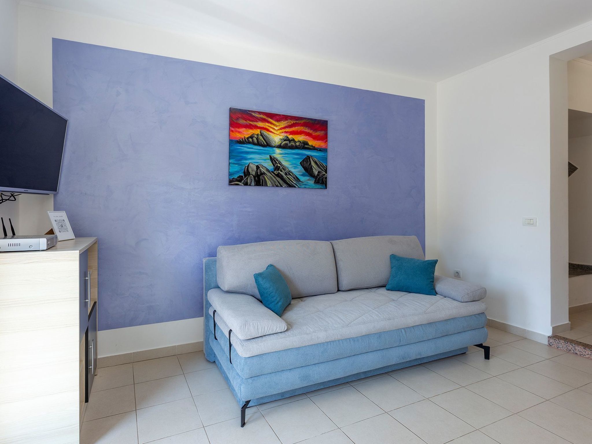 Un salon avec un canapé bleu clair, un tableau représentant un coucher de soleil sur des rochers et un téléviseur à écran plat posé sur un meuble en bois.