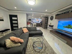 Ein modernes Wohnzimmer mit einem Ecksofa, einem Flachbildfernseher und einer Küche im Hintergrund.