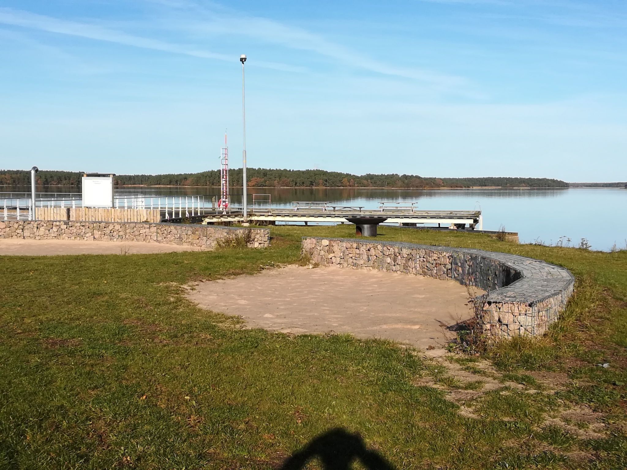 A lakeside park with a stone wall, a metal fence, and a dock extending into the water.