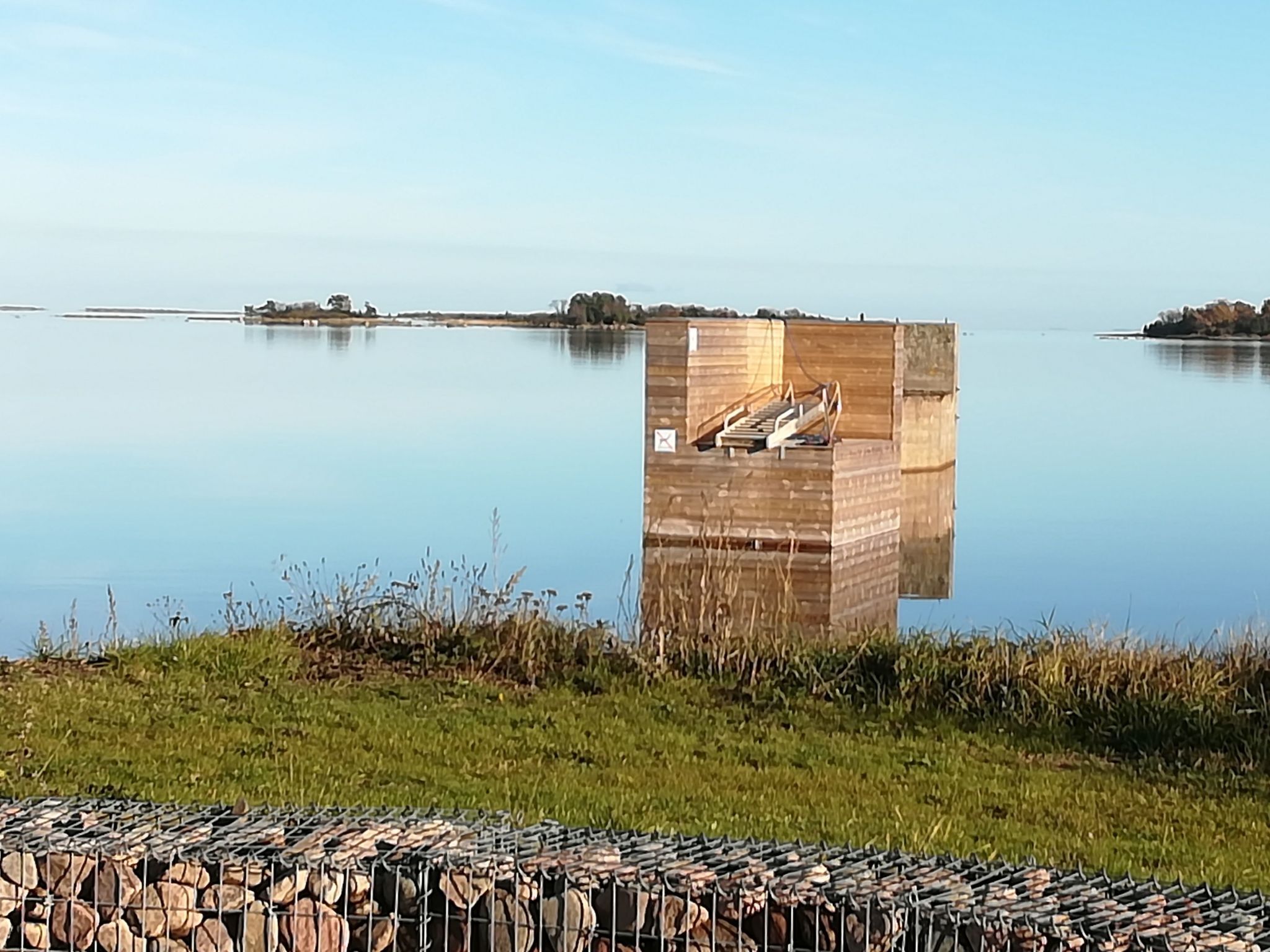 Une structure en bois avec un escalier menant à une plateforme, entourée d’une étendue d’eau calme et de zones verdoyantes.