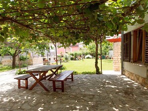 Un patio couvert avec une table en bois et des bancs, entouré de verdure et doté d’un sol en pierre.