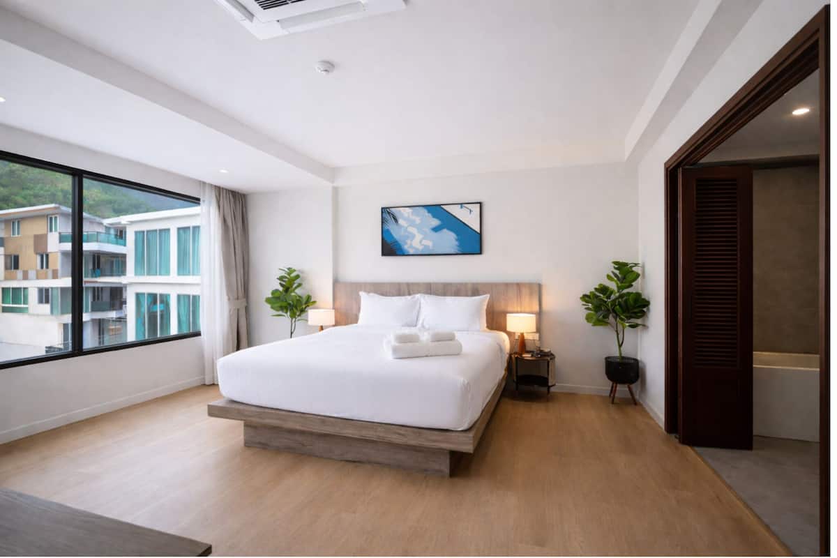 A modern hotel room with a large bed, a view of buildings through the window, and a painting on the wall.