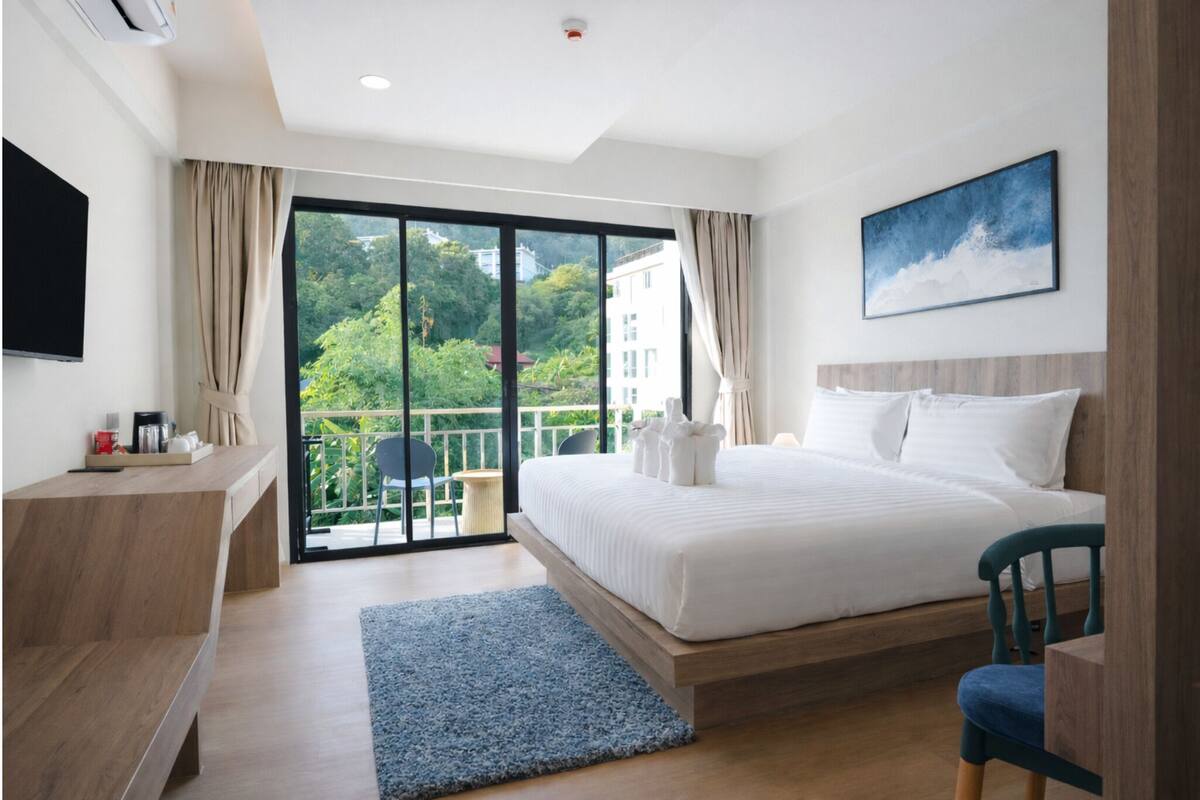 A modern hotel room with a large bed, a desk, a chair, and a balcony with a view of greenery and buildings.