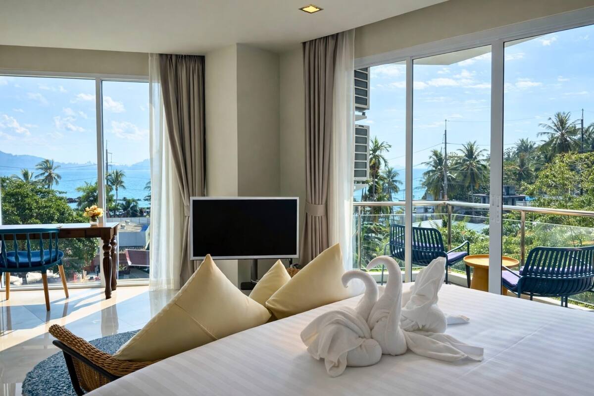 A hotel room with a large bed, a flat-screen TV, a balcony with chairs, and a view of the ocean and palm trees.