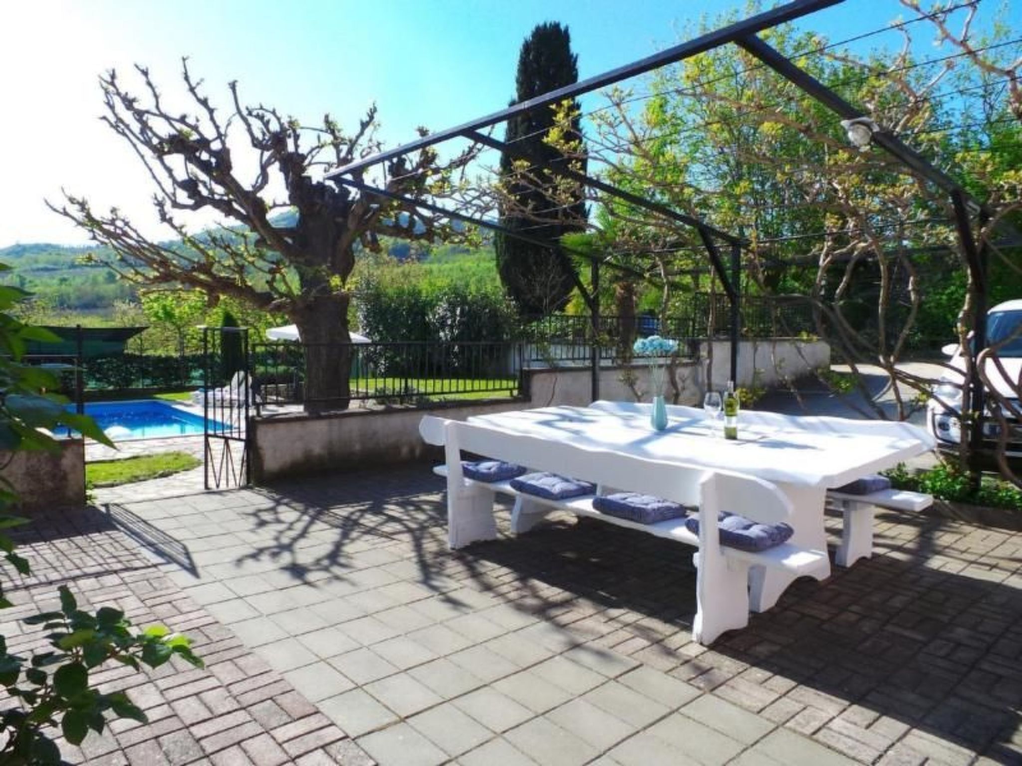 A patio with a white table and benches, surrounded by a pool and greenery.