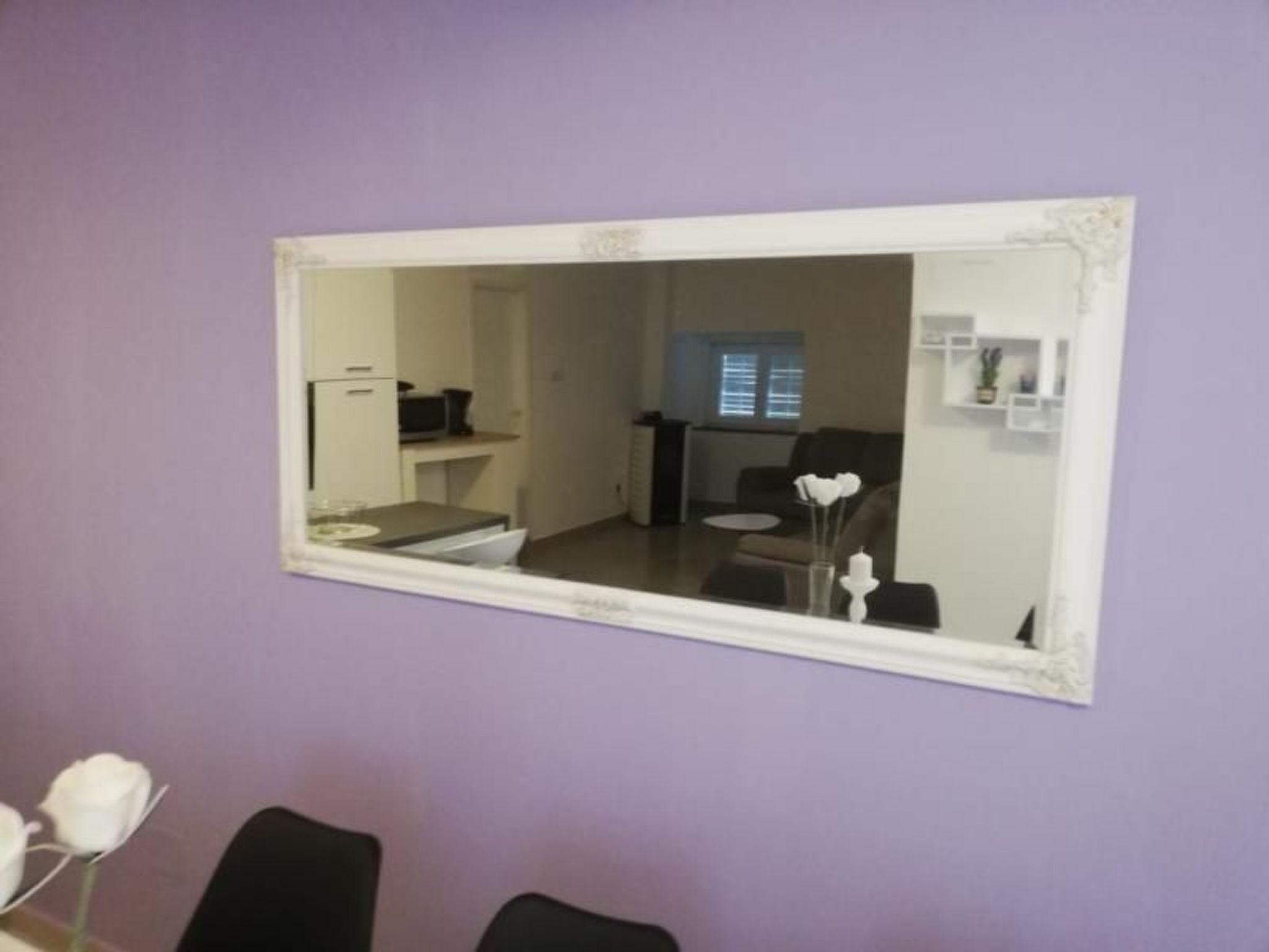 A room with a large mirror on a purple wall, a dining table with chairs, and a kitchen area visible through the mirror.