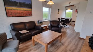 A modern living room with a wooden coffee table, leather sofa, and armchair. There is a dining area with a table and chairs, and a large window providing a view of a field. - Gemütliches Ferienhaus De Blenck by Interhome (Callantsoog)