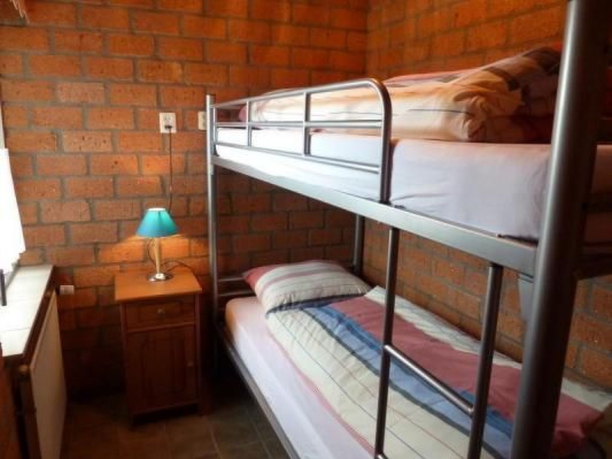 Une chambre avec un lit superposé, un mur en briques, une petite table de chevet en bois avec une lampe et un bureau.