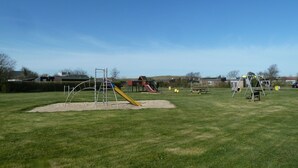 A playground with various play structures, including a slide and swings, set in a grassy field with buildings in the background. - Gemütliches Ferienhaus De Blenck by Interhome (Callantsoog)