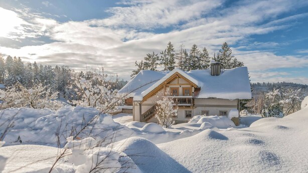 Une maison recouverte de neige, avec une façade en bois et un balcon, entourée d’arbres et de neige.