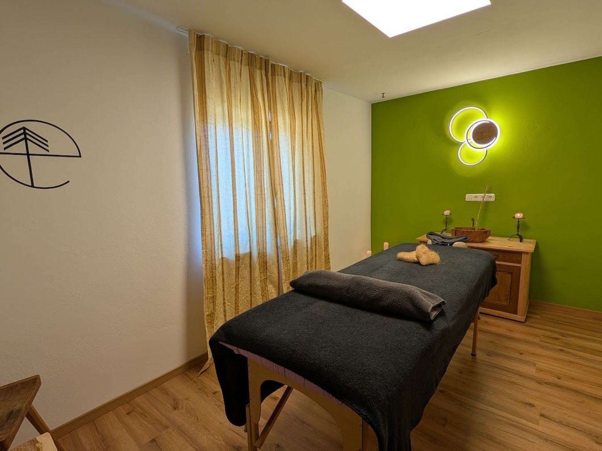 A room with a massage table, wooden floor, green wall, and a window with curtains.