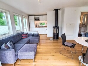 A modern living space with a sofa, ottoman, dining table, chairs, a kitchen area, and a wood-burning stove. - Ljugarn 2 by Interhome (Ljugarn)