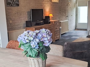 A room with a brick wall, a television on a wooden stand, a lamp, a wicker basket with flowers, and a door leading outside. - Gemütlicher Bungalow für 4 Personen by Interhome (Callantsoog)