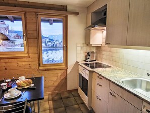 Een compacte keuken met houten wanden, een raam met uitzicht op een besneeuwd landschap, een kleine eettafel met ontbijtartikelen en een inbouwoven.