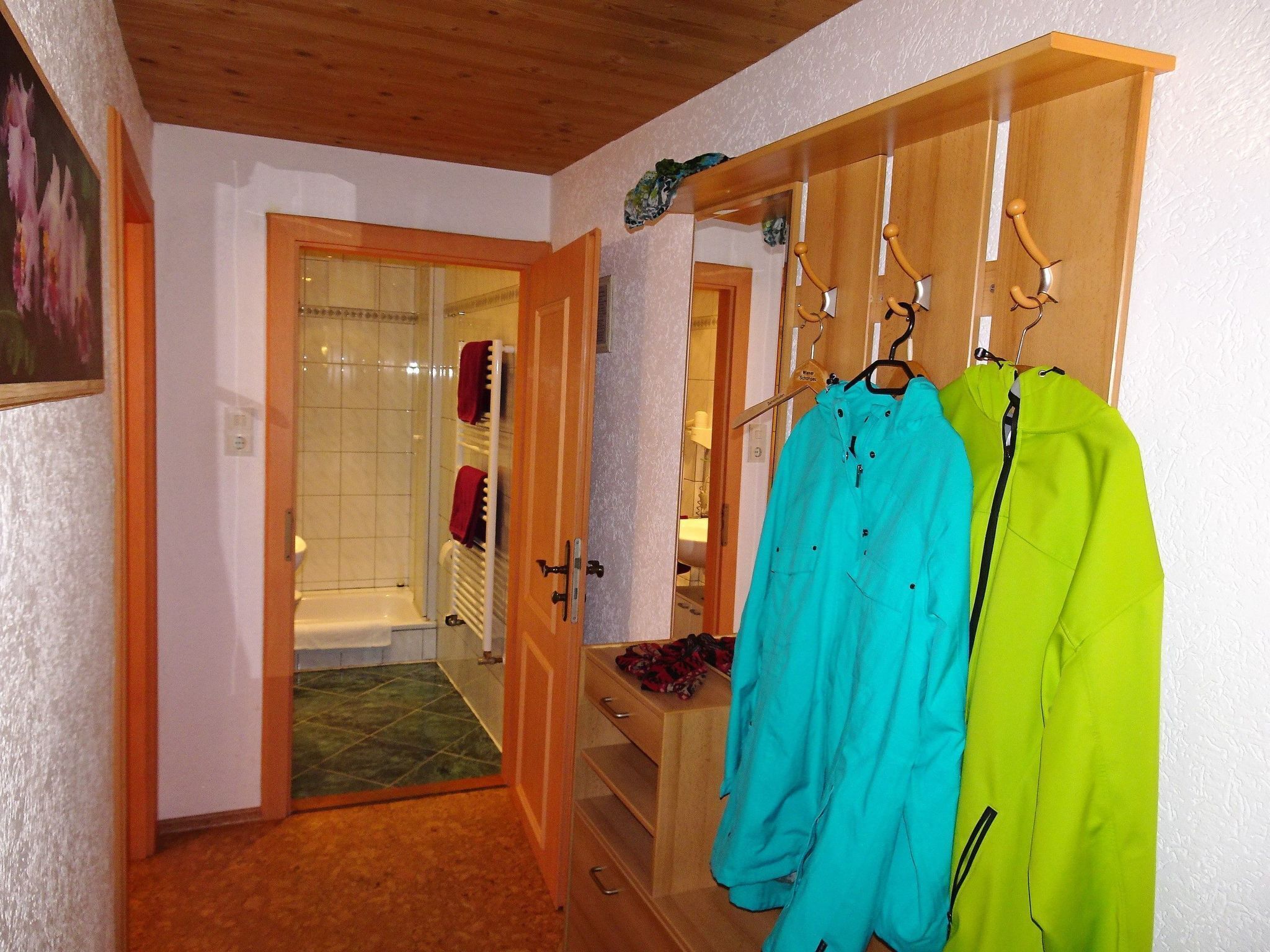 Un couloir avec une porte en bois donnant sur une salle de bain, un porte-manteau avec deux vestes, et une petite table sur laquelle est posée une serviette rouge.