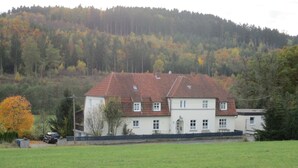 Une maison à deux étages, avec un toit recouvert de tuiles rouges, entourée d’une pelouse verdoyante et bordée, au loin, par une colline boisée.