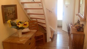Un escalier en bois menant à un deuxième étage, une commode en bois ornée d’un vase de tournesols et un miroir accroché au mur.
