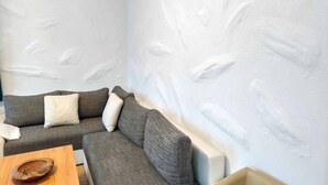 Een hoek van een kamer met een grijze bank, een houten bijzettafeltje en een witte muur met een structuur.