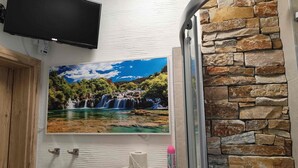 Een badkamer met een wandtelevisie, een waterval schilderij, een douche met een stenen muur en toiletartikelen op het aanrecht.