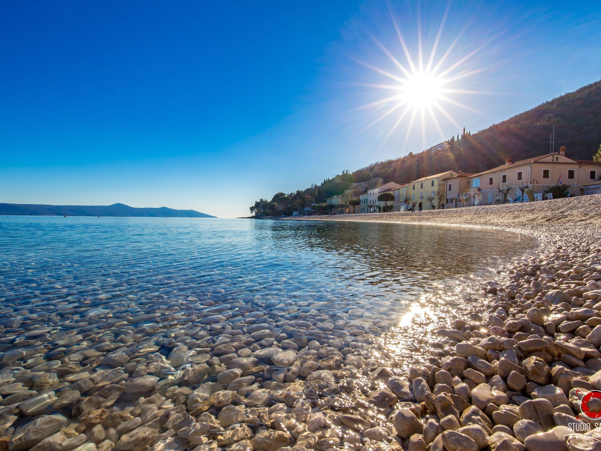 A pebble beach with clear water, a sunlit sky, and buildings along the shore.