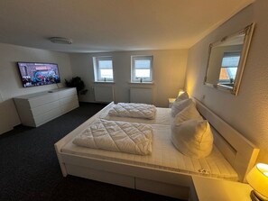 Ein Schlafzimmer mit Bett, Fernseher, Kommode und zwei Fenstern.