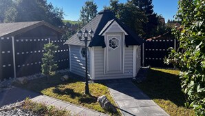 Eine kleine Holzhütte mit schwarzem Dach und einem dekorativen Fenster, umgeben von einem Garten mit einem Kiesweg und einem Holzzaun.