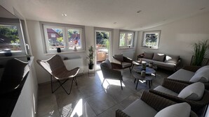Ein sonnendurchflutetes Zimmer mit Korbmöbeln, großen Fenstern und Blick ins Freie.