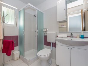 Ein Badezimmer mit Duschkabine, weißer Toilette, Waschbecken mit Spiegel und einer kleinen Stehocker.